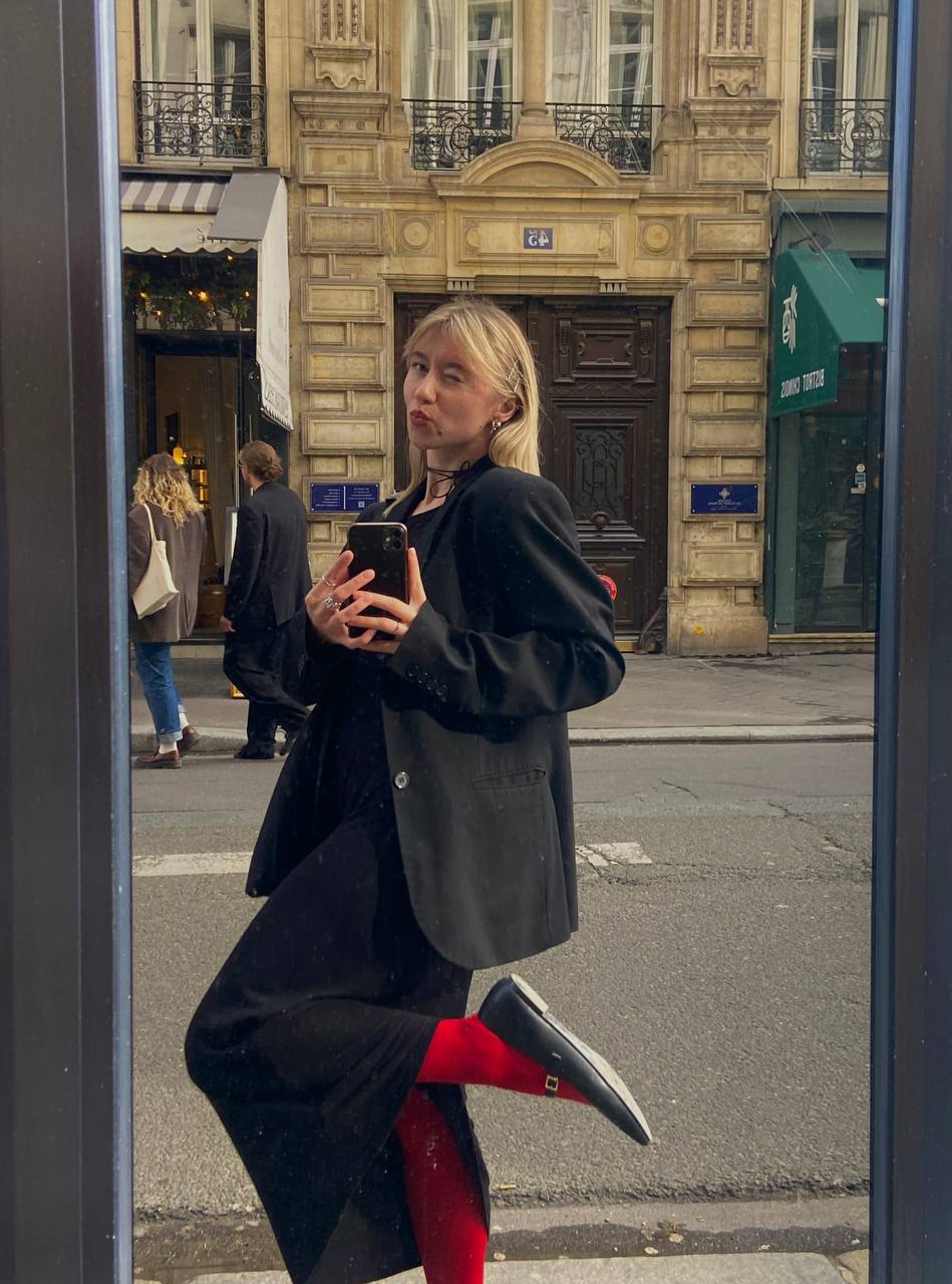 Девушка делает селфи в зеркале на улице в Париже. На ней черный оверзайз пиджак, черное платье, красные колготки и черные балетки. На шее – чокер.